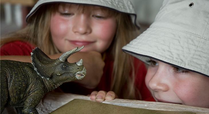 Two smiling children look at a Triceratops toy.